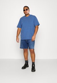 Camiseta de algodón azul combinada con pantalones cortos de mezclilla azul, complementada con botas negras hasta el tobillo y gafas de sol cuadradas oscuras. Atuendo simple y casual.