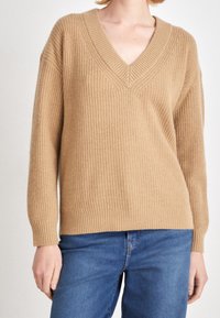 Pull beige en maille avec un col en V profond, des poignets et un ourlet côtelés, présentant une coupe décontractée et un tissu texturé. Porté sur un jean bleu.