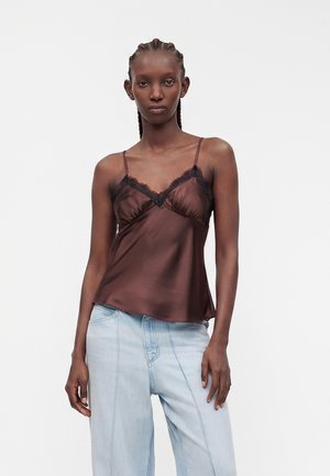 Femme aux cheveux tressés portant un caraco en satin marron avec une bordure en dentelle noire et un jean bleu clair taille haute à jambes larges.
