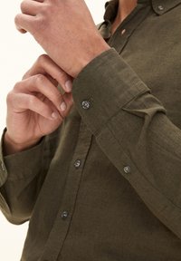 Olijfgroene button-up shirt van textuurstof, met een gebogen kraag en verstelbare knoopmanchetten, voorzien van grijze knopen en zichtbare stiksels.