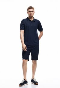 Marineblauwe polo met korte mouwen, gecombineerd met marineblauwe shorts. Model draagt marineblauwe en witte sneakers. Soepele textuur, casual pasvorm.
