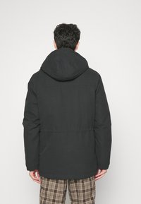 Black padded jacket with a hood, featuring a cinched waist, elastic cuffs, and a smooth texture. Worn over checked brown pants.