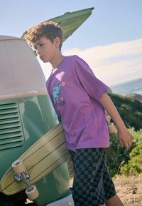 Paars T-shirt met korte mouwen met een grafisch ontwerp en tekst op de voorkant, gecombineerd met zwart-wit geruite shorts. Houdt een lichtgekleurde skateboard vast.