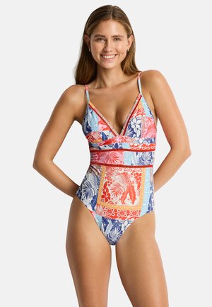 Donna sorridente che indossa un costume intero colorato con motivi floreali e geometrici e sottili bretelle su uno sfondo semplice.