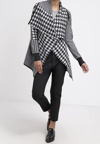 Šátek s černobílým houndstooth vzorem a asymetrickým designem, kombinovaný s černými kalhotami a plochými botami na klouzání.