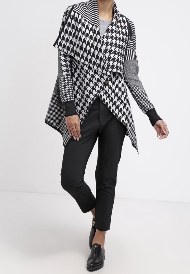 Šátek s černobílým houndstooth vzorem a asymetrickým designem, kombinovaný s černými kalhotami a plochými botami na klouzání.