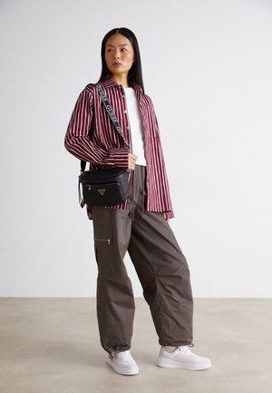 Vrouw staand met een bordeauxrode en witte gestreepte blouse, wit topje, bruine cargobroek, witte sneakers en een zwarte schoudertas.