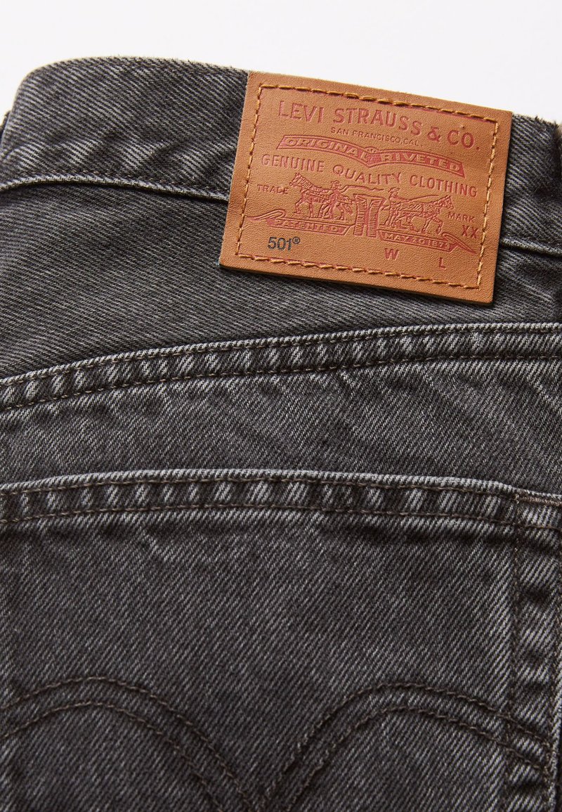 Les jeans Levi's 501 sont en denim gris foncé, avec une patch en cuir au niveau de la taille portant un logo embossé, et des coutures incurvées distinctives sur les poches arrière.