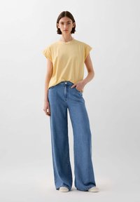 Gelbes T-Shirt mit kurzen Ärmeln aus weichem Stoff, kombiniert mit weit geschnittenen blauen Jeans aus Denim. Die Jeans verfügen über einen klassischen Knopfverschluss und Taschen.