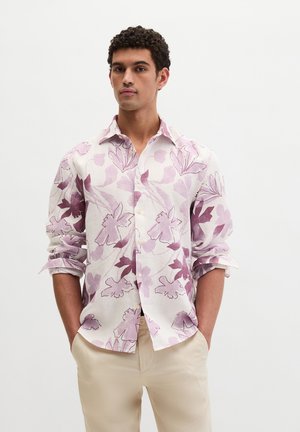 Mann trägt ein langärmeliges weißes Hemd mit lila Blumenmuster und beigefarbene Hose, steht mit Händen in den Taschen vor einem einfarbigen Hintergrund.