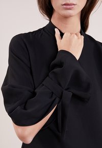 Blouse noire à col montant, avec des manches volumineuses nouées au niveau des poignets. Tissu lisse et fluide créant un look minimaliste.