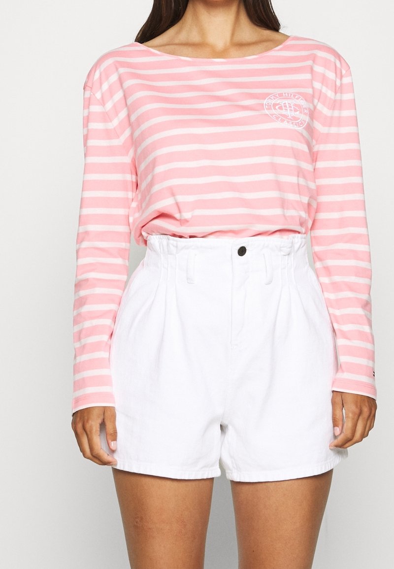T-shirt a maniche lunghe a righe rosa e bianche abbinato a pantaloni corti bianchi a vita alta. Il T-shirt presenta un piccolo logo sul lato sinistro del petto.