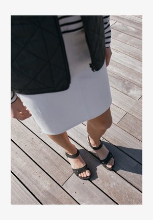 Sandales noires à bout ouvert, avec une sangle de cheville réglable et un dessus texturé, associées à une jupe blanche mi-longue et un haut rayé.