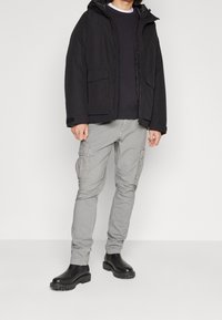 Svart huva jacka med framfickor, lager på lager över en mörk tröja. Ljusgrå cargobyxor med flera fickor. Svarta ankelstövlar.