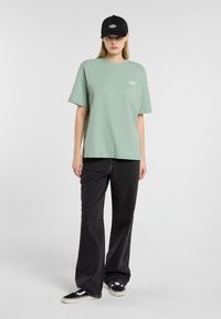 T-shirt oversize vert menthe à manches courtes, associé à un jean noir à jambes larges et des baskets noires, agrémenté d'une casquette noire.