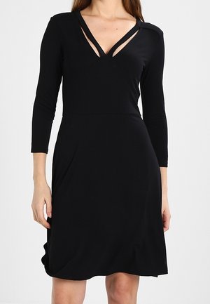 Femme portant une robe noire longueur genou avec des manches trois-quarts et un décolleté en V doté de détails à double bretelle.