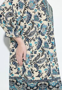 Mano descansando sobre un vestido beige con patrones florales y de paisley en azul y negro, con el puño elástico visible en la manga de tres cuartos.