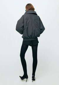 Puchowa czarna kurtka z wysokim kołnierzem, elastycznym dołem i opuszczonymi ramionami, zestawiona z czarnymi rajstopami i szpilkami.