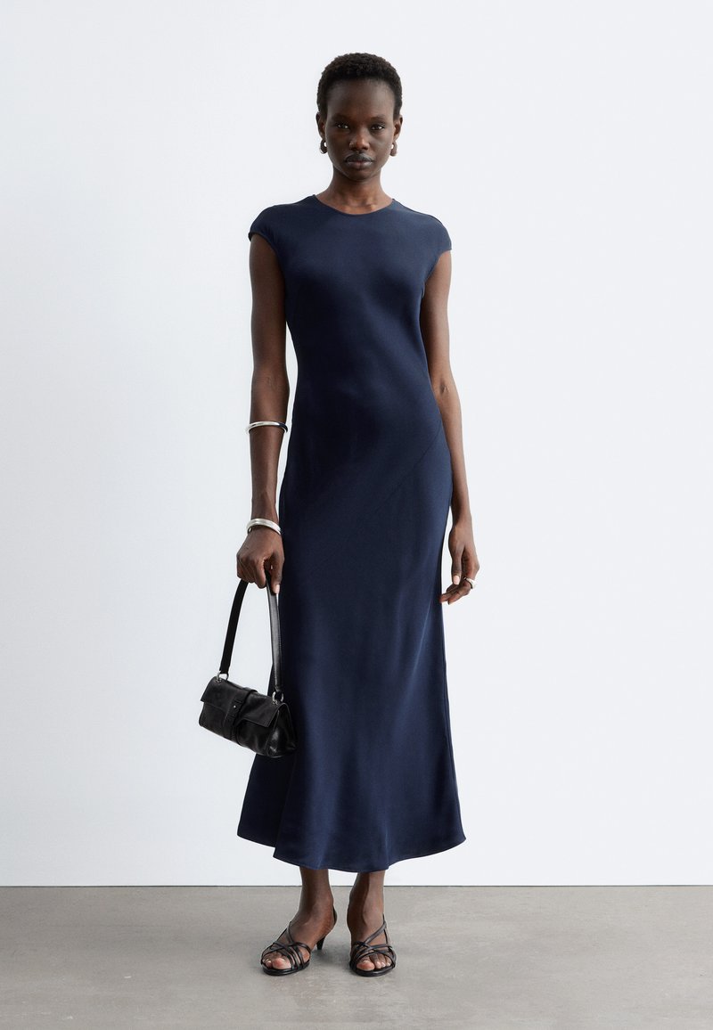 Femme debout portant une robe midi bleu marine à manches courtes, tenant un petit sac à main noir, avec des bracelets argentés et des sandales noires à brides.