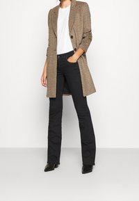 Blazer largo de cuadro marrón sobre una camisa blanca, combinado con jeans negros de campana y botines negros. Diseño simple y estructurado.