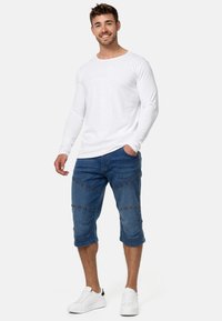 Cămașă albă cu mânecă lungă și pantaloni scurți din denim albastru, până la genunchi, cu cusături în unghi. Design simplu, purtat cu adidași albi.