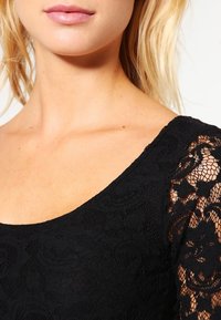 Svart spets-topp med blommönster, med rundad halsringning och genomskinliga spetsärmar. Den släta texturen kontrasterar mot den intrikata detaljerna.