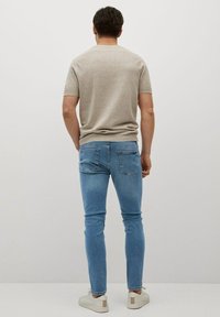 Suéter beige de manga corta, jeans ajustados azul claro y zapatillas blancas con acentos marrones, visto desde la parte posterior contra un fondo liso.