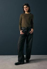 Dunkelgrünes Langarmoberteil, locker sitzende schwarze Denim-Jeans, schwarze Schuhe, getragen von einem Model vor einem dunklen Hintergrund, minimalistisches Design.