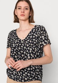 Top preto de mangas curtas com decote em V, apresentando um padrão floral branco e um tecido suave e leve. Ajuste casual, com um leve caimento.