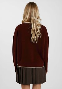 Maglione bordeaux lavorato a maglia con vestibilità comoda e orlo a coste, abbinato a una gonna plissettata a quadri marrone. La texture sembra morbida e accogliente.
