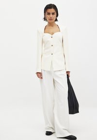Balta šūta žakete ar kvadrātveida dekoltē, zelta pogām un garām piedurknēm, apvienota ar baltām platām biksēm un melnu rokassomu.