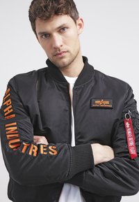 Jachetă bomber neagră cu broderie portocalie "ALPHA INDUSTRIES" pe mâneci și patch cu logo, având manșete ribbate și un tag roșu "REMOVE BEFORE FLIGHT".