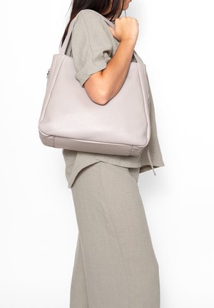 Vrouw in beige outfit draagt een grote lichtgrijze leren handtas op haar schouder tegen een witte achtergrond.