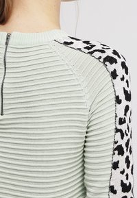 Hellgrüner Rippstrickpullover mit einem schwarz-weißen Leopardenmuster an den Schultern und einem Reißverschluss auf der Rückseite.