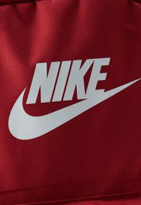 Λευκό λογότυπο Nike και σύμβολο swoosh σε υφασμάτινη επιφάνεια κόκκινου χρώματος με υφή, πιθανόν μέρος τσάντας ή ένδυσης.