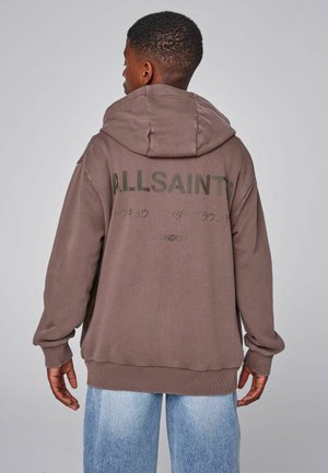 Young person wearing a brown AllSaints hoodie with text and Japanese characters on the back, paired with light blue jeans.