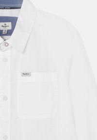 Biała koszula z guzikami z pojedynczą kieszenią na klatce piersiowej, niebieskim podszyciem kołnierzyka i małą metką z logo "Pepe Jeans" na kieszeni. Gładka tekstura.