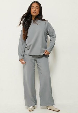 Femme aux longs cheveux foncés portant un haut gris ample à manches longues et un pantalon large avec des baskets blanches, debout devant un fond clair.