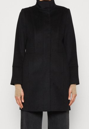 Femme portant un manteau noir à col montant avec des manches longues, assorti à des gants beiges sans doigts et un pantalon foncé, debout devant un fond uni.