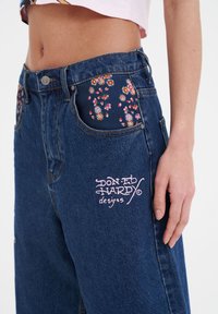 Vaqueros de mezclilla con un lavado azul oscuro, forros de bolsillo con un patrón floral y un logo bordado en rosa en el bolsillo frontal.