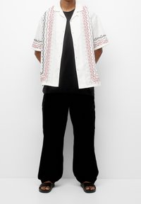 Kortærmet hvid skjorte med rød og sort geometrisk broderi, parret med sorte bukser og sorte sandaler. Afslappet pasform og afslappet design.