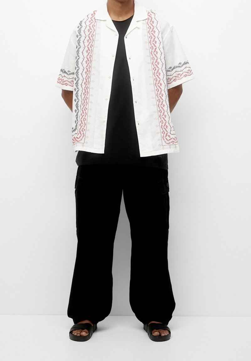 Kortærmet hvid skjorte med rød og sort geometrisk broderi, parret med sorte bukser og sorte sandaler. Afslappet pasform og afslappet design.