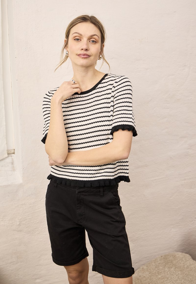Jeune femme aux cheveux clairs portant un haut à manches courtes rayé noir et blanc et un short noir jusqu'aux genoux, posant à l'intérieur contre un mur clair.