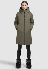 Manteau long matelassé vert olive avec fermeture éclair, capuche surdimensionnée, poches latérales et accents noirs. Associé à un legging noir et des bottes.
