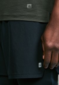 Grey-green athletic shirt with a smooth texture and a small silver logo. Paired with dark shorts featuring a similar logo at the hem.