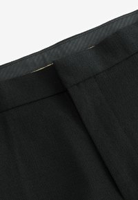 Nahaufnahme eines schwarzen Stoffes, der eine strukturierte Oberfläche mit diagonalem Gewebe zeigt. Verfügt über einen glatten Bund und eine kontrastierende Futter in einem subtilen Muster.