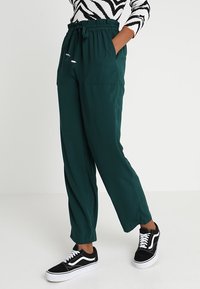 Pantalons amples de couleur vert foncé avec une taille élastique, des cordons de serrage, et deux poches latérales, associés à des baskets noires et blanches.