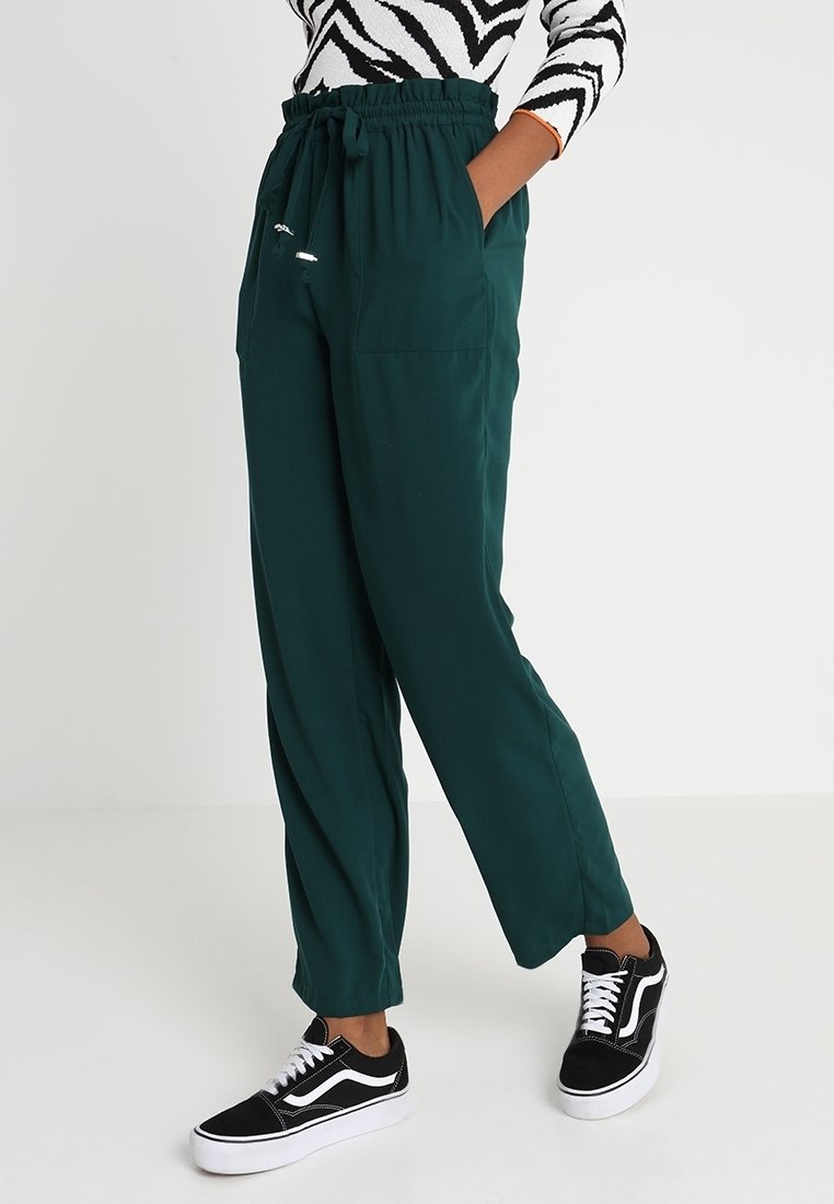 Pantalons amples de couleur vert foncé avec une taille élastique, des cordons de serrage, et deux poches latérales, associés à des baskets noires et blanches.