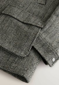 Pelēks zigzag audums ar maigu tekstūru, kas izceļas ar slāņotu dizainu un sudraba spiedpogu detaļu.