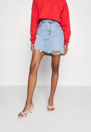Jupe en denim mini bleu clair avec un ourlet effiloché, conçue avec une taille ajustée et des détails usés, assortie à des talons blancs à lanières.